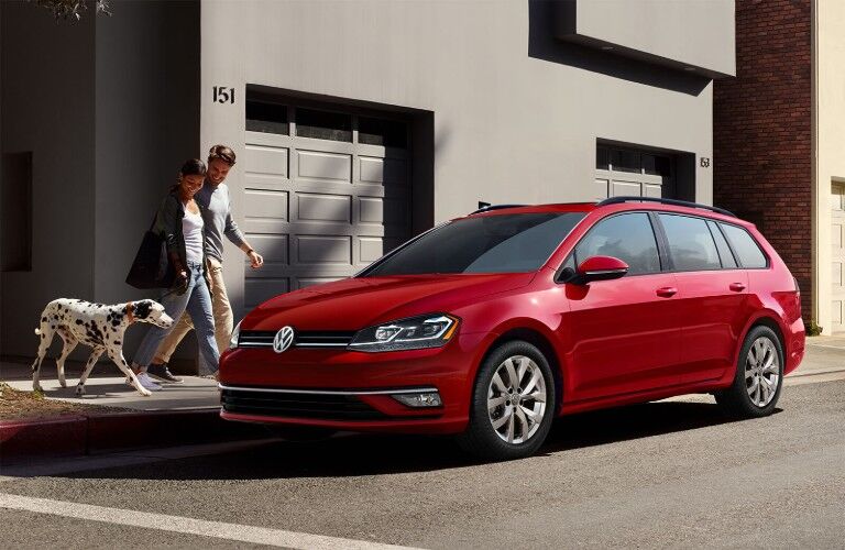 Front driver angle of a red 2019 Volkswagen Golf SportWagen parked on the side of a street with a couple and their dalmatian walking towards it
