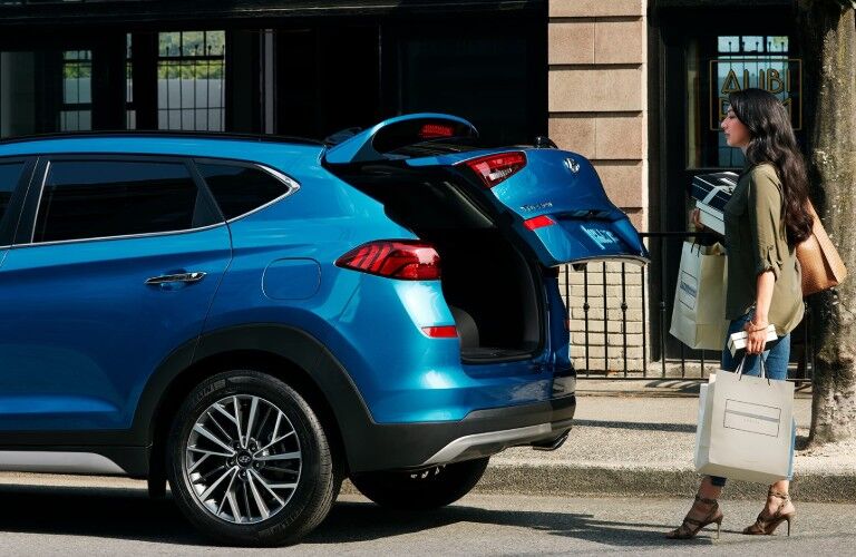 Rear driver angle of a blue 2020 Hyundai Tucson with its liftgate opening and a woman standing behind with bags