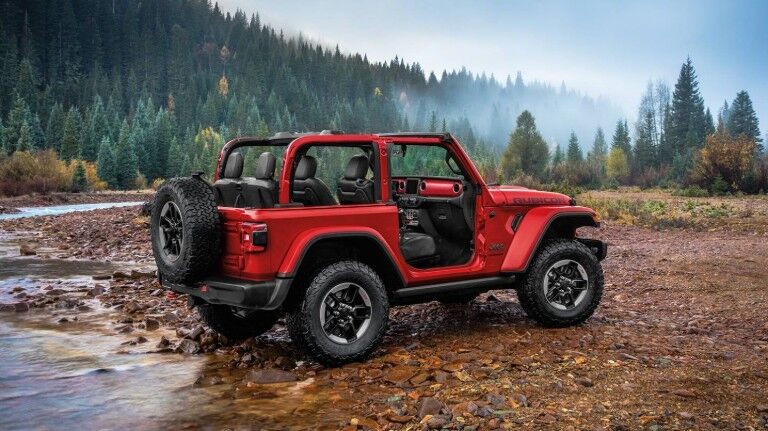 2020 Jeep Wrangler exterior side shot with firecracker red paint color and doors removed on wet earth and wood chips near a lake and forest with a foggy sky