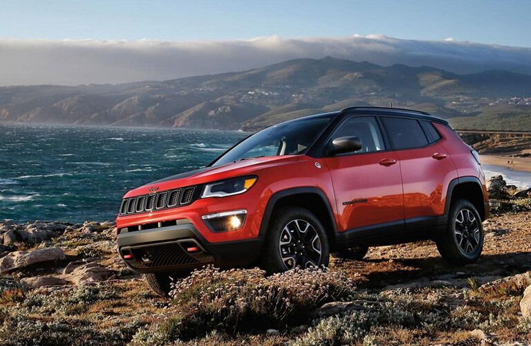 2020 Jeep Compass parked on a rocky and flower covered beach near the blue sea and drifting mountains