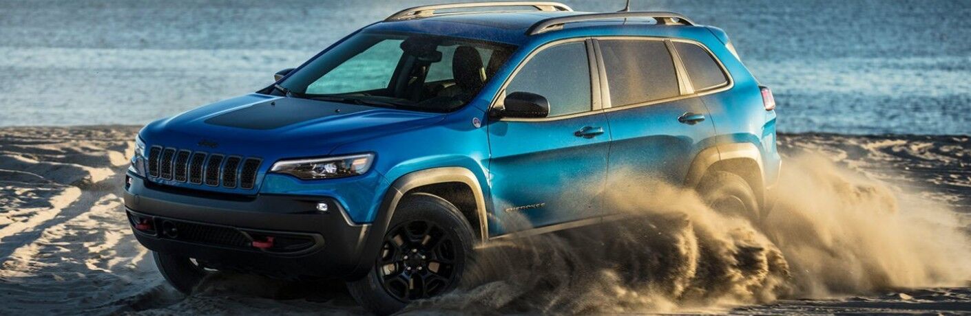 2020 Jeep Cherokee exterior side shot with Hydro Blue Pearl Coat paint color driving on a beach as it kicks up sand with an ocean wave background