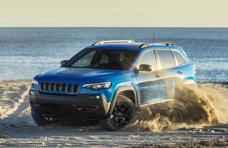 2020 Jeep Cherokee exterior shot with blue paint color kicking up dust clouds as it drives on a sandy beach near the ocean