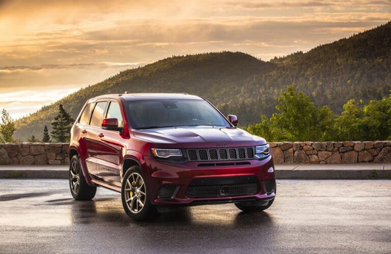 2020 Jeep Grand Cherokee exterior front shot with dark red paint color parked on a wet lot near grassy mountains as the sun sets in the sky
