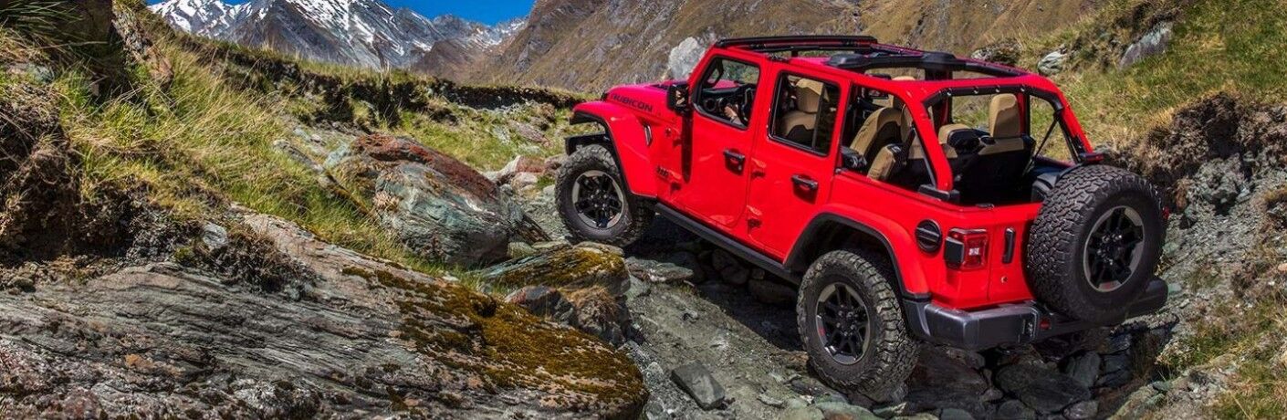 2020 Jeep Wrangler Rubicon exterior shot with red paint color climbing up a rocky grass covered cliff near blue skies and snow capped mountains
