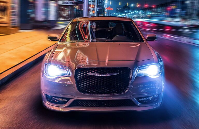 2019 Chrysler 300 exterior front shot with white paint color and headlights on driving through a busy city at night
