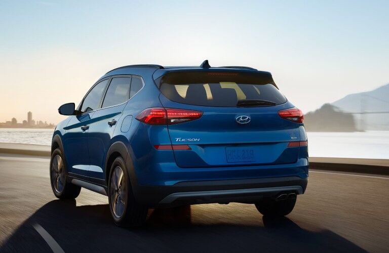 Rear driver angle of a blue 2020 Hyundai Tucson driving by a lake with a city in the background